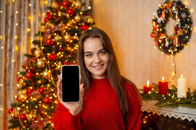 Happy young woman standing in front of christmas tree shows the smartphone to the camera Winter holidays getting messages and communication online concept copy space