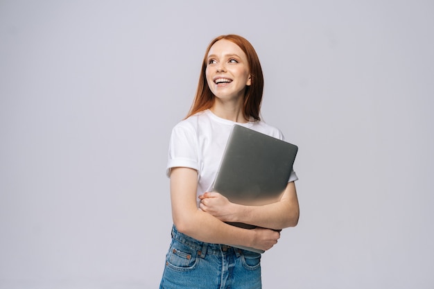 Happy young woman student holding laptop computer and looking away on isolated gray background