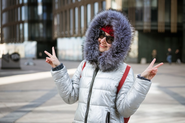 Happy young woman in sunglasses in a winter jacket with a backpack near the building. Travel concept