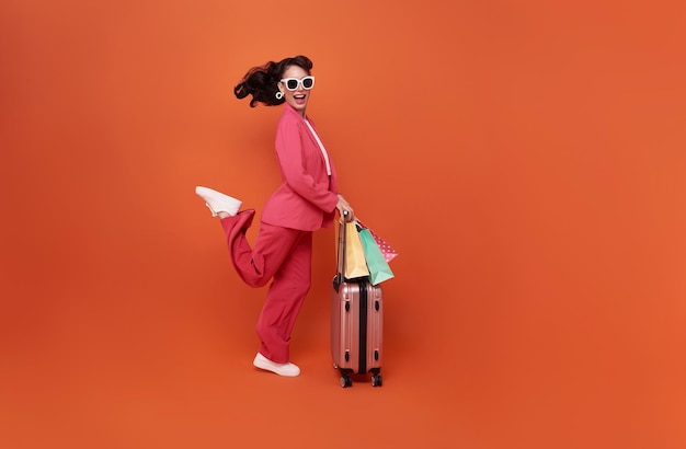 Happy young woman traveler drag luggage and shopping bag isolated on orange copy space background