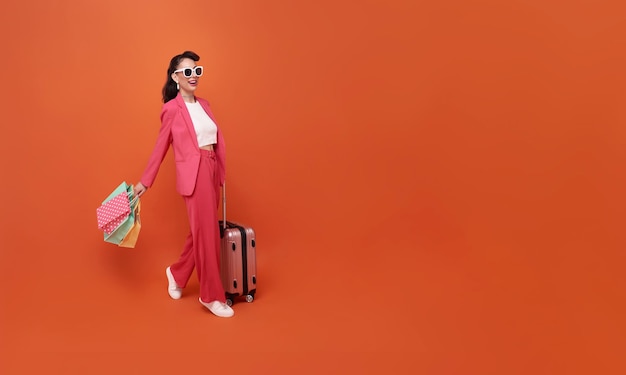 Happy young woman traveler drag luggage and shopping bag isolated on orange copy space