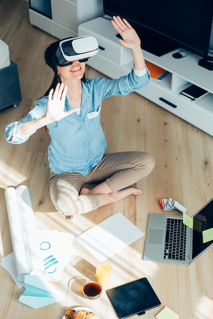 Happy young woman in virtual reality glasses sitting on the floor with a laptop and stationery supplies by her side and putting head and hands up