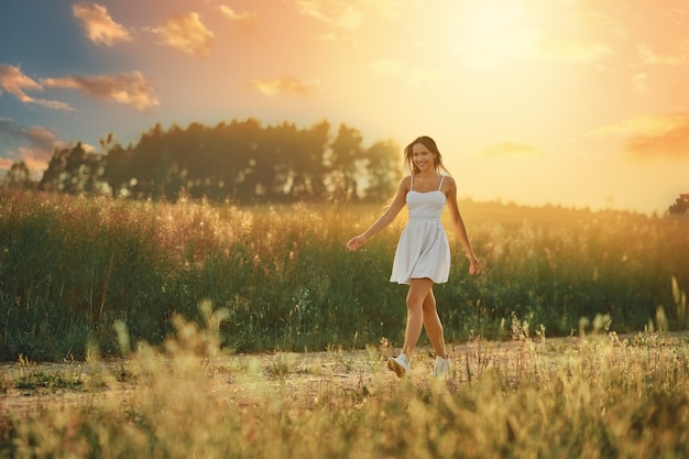 Happy young woman walking along country road in the meadow during sunset