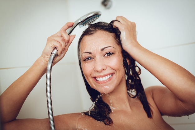 Happy young woman washing face and hair while showering under shower head.