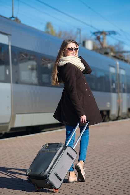 Happy young woman wearing glasses dressed in coat with big travel bag at the railway station on the train background