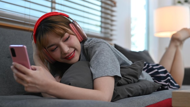 Happy young woman wearing headphone lying on couch and using mobile phone spending leisure weekend time at home