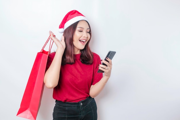 A happy young woman wearing Santa Claus39 hat is smiling while holding Christmas presents and her smartphone