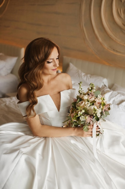 Happy young woman in a wedding dress enjoying with a bridal bouquet on the bed