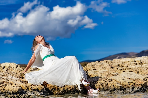 Happy young woman in white dress sitting on the sand rock and enjoying on elafonisi beach