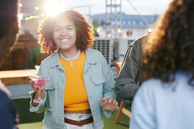 Happy young woman with cocktail looking at one of friends while enjoying party