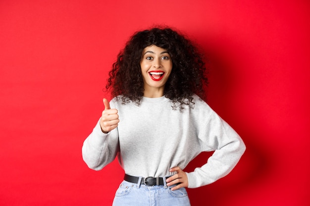 Happy young woman with curly hair praising good work, say well done and show thumb up gesture, approve and praise you, standing on red wall