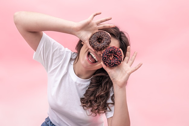 Happy young woman with donuts in her hands smile, holds pink donuts near her eyes her hands, pink background.