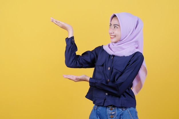 Happy young woman with happy facial expression shows something in her hand wearing hijab and casual clothes in yellow