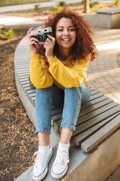 Happy young woman with red curly hair sitting on a bench at the park, taking pictures with photo camera