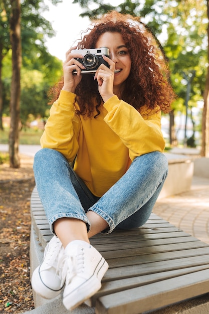 Happy young woman with red curly hair sitting on a bench at the park, taking pictures with photo camera