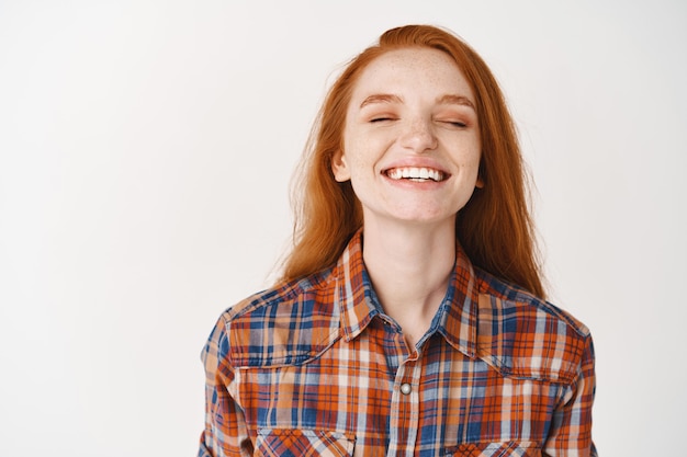 Happy young woman with red hair, close eyes and smiling dreamy, standing over white wall