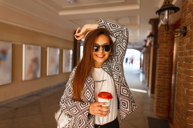 Happy young woman with a smile in sunglasses and a fashionable jacket drinks coffee and smiles, walks in the city