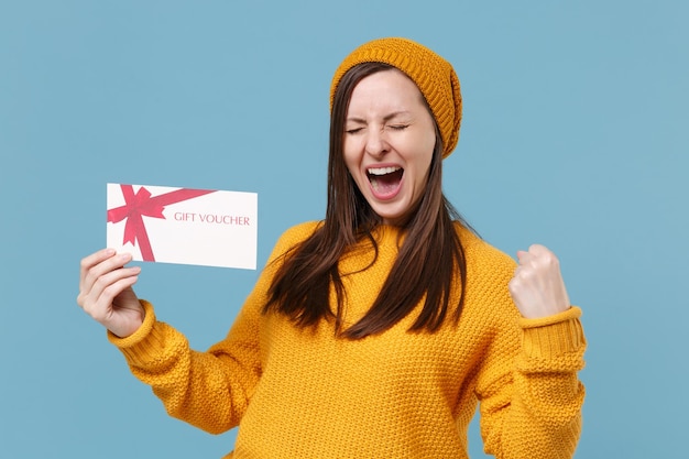 Happy young woman in yellow sweater hat posing isolated on blue background studio portrait. People sincere emotions lifestyle concept. Mock up copy space. Doing winner gesture hold gift certificate.