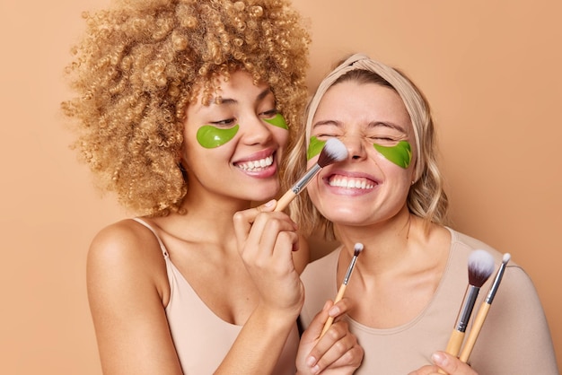 Happy young women apply beauty patches use cosmetic brushes smile gladfully undergo facial treatments stand next to each other isolated over beige background Wellness and cosmetology concept