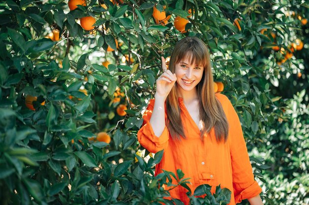 Hapy young girl in orange dress is pointing up with forefinger by looking at camera in orange garden