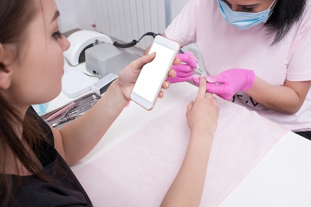 Hardware manicure. Smartphone in hands of client. Manicure master doing beauty procedure, girl chating on phone