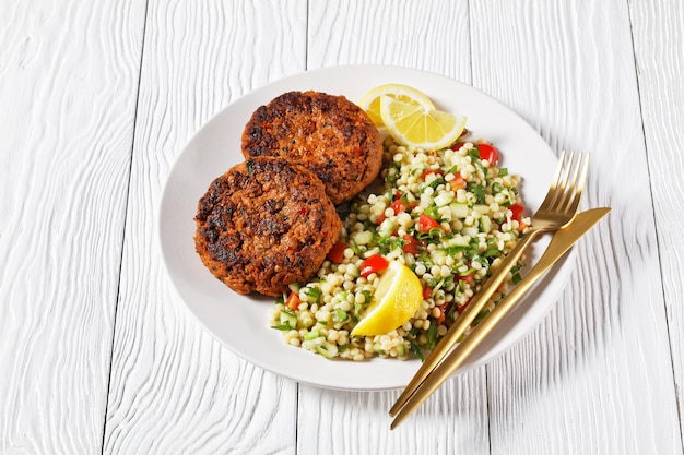 Harissa lamb burgers served with pearl couscous salad on a plate with lemon slices on a white wooden table with golden cutlery, horizontal view from above