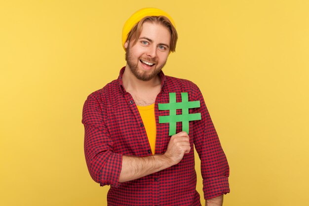 Hashtag, internet trends. Positive hipster guy in checkered shirt smiling and holding hash sign on his chest, symbol of viral message social network. indoor studio shot isolated on yellow background