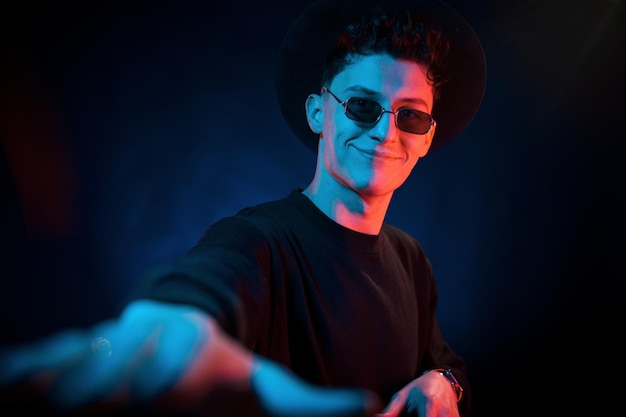 In hat and glasses Neon lighting Young european man is in the dark studio