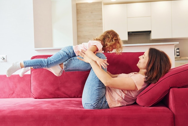 Having fun on red sofa Young mother with her little daughter in casual clothes together indoors at home