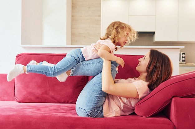 Having fun on red sofa Young mother with her little daughter in casual clothes together indoors at home