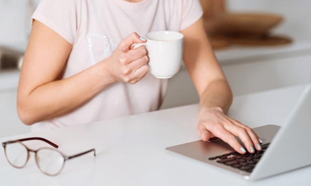 Having some plans. Delicate bright elegant woman using her computer finding required ingredients while having a breakfast and sitting in sunlit kitchen