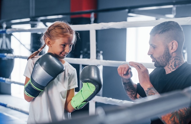 Photo having sparring with each other on the boxing ring. young tattooed boxing coach teaches cute little girl in the gym.