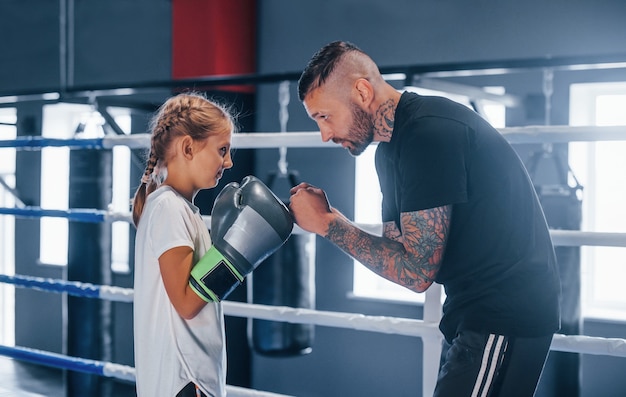 Having sparring with each other on the boxing ring. Young tattooed boxing coach teaches cute little girl in the gym.