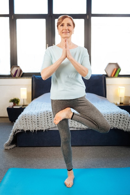 Having yoga session. Smiling light-haired lady wearing comfortable outfit during yoga training while standing in bedroom on mat