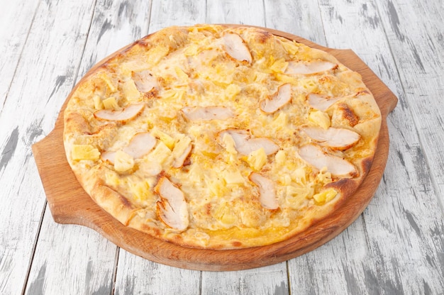 Hawaiian pizza with chicken fillet, pineapple and mozzarella cheese. On a round wooden board. Light wooden background. View from above.