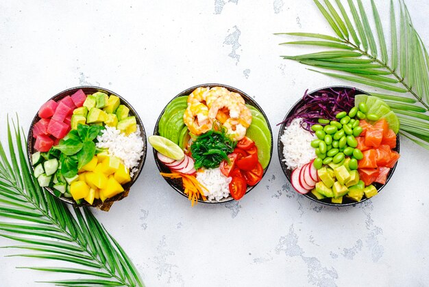 Hawaiian poke bowl set tuna salmon shrimp with avocado mango radish rice and other ingredients Soy sauce and sesame dressing White table background palm leaves top view