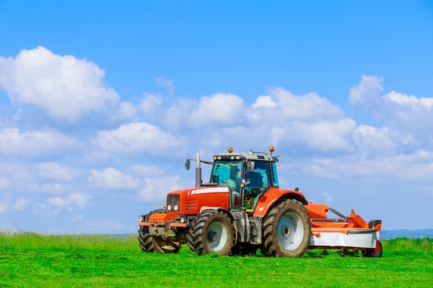 Haymaking. A large red tractor with a mower mows the grass in the field on a sunny day.