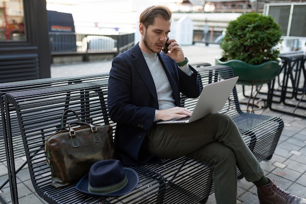 The head of a middleaged man works on a laptop and speaks on a mobile phone while sitting on a bench