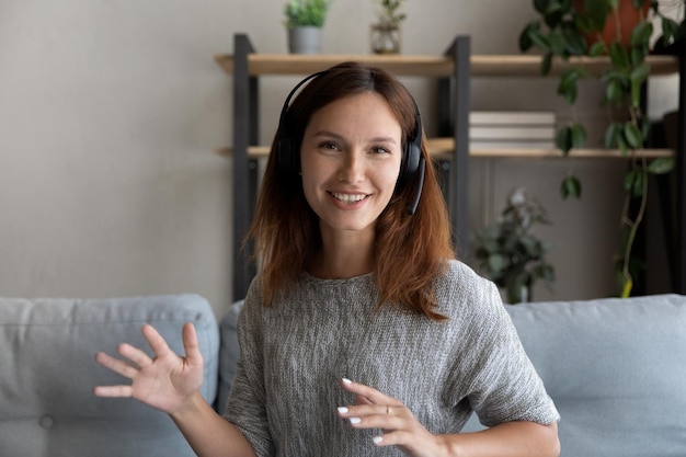 Photo head shot portrait of smiling woman in headphones speaking and looking at camera sitting on couch at home teacher mentor recording webinar leading online lesson involved in internet meeting