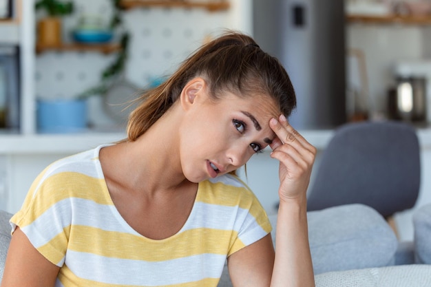 Headache Closeup photo of a young woman who is sitting on a sofa touching her head while suffering from a migraine