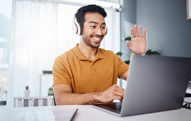 Headphones laptop and man on video call webinar or virtual meeting talking and online in work from home office Happy Asian person waves hello on his computer pc in client chat for tech business