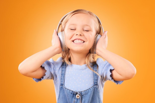 Headphones smile or child streaming music to relax with freedom in studio on orange background Face singing or happy girl singer listening to a radio song sound or audio on an online subscription