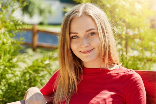 Headshot of beautiful female with freckled face and warm blue eyes smiling gently into camera wearing red sweater resting in park at bench. Cheerful student girl having break in open air after classes