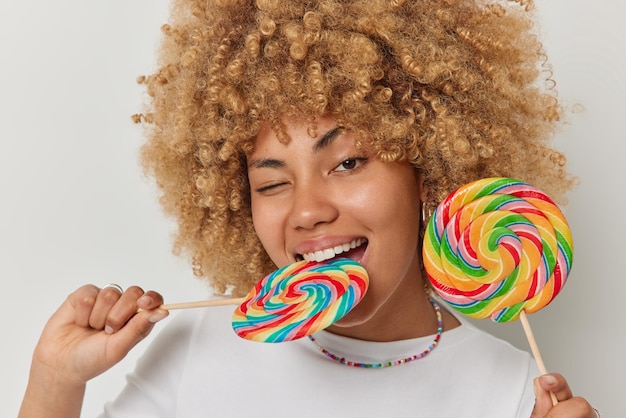 Headshot of curly haired beautiful woman bites multicolored caramel candy on stick winks eye has glad expression enjoys eating delicious sweet rainbow lollipops poses against white background