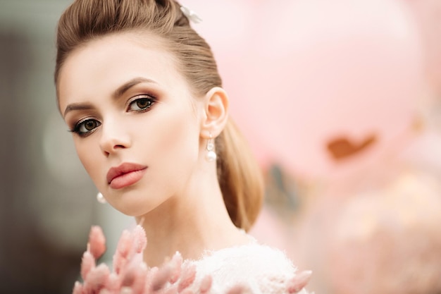 Headshot of gorgeous brunette woman with flawless skin high hairdo professional make up and bunch of pink flowers looking at camera Unfocused pink background