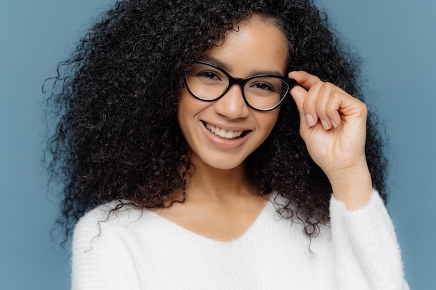 Headshot of optimisitc young African American woman with crisp hair holds hand on frame of glasses wears white sweater being in good mood after successful day isolated over blue background