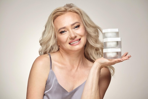 Headshot portrait of mature woman holding jars of cream for skin care on grey isolated background