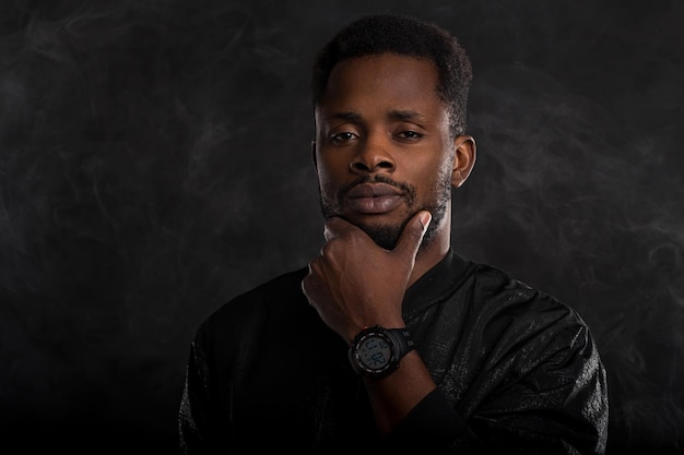 Headshot of serious dark skinned male holding chin looking mysteriously at camera, dressed in black windbreaker jacket, wristwatces, standing against dark background with white smoke. Close up.