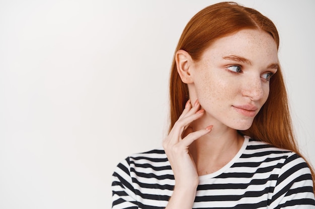 Headshot of tender young woman with pale skin, red hair and blue eyes, turn head right and gently touching perfect face with natural makeup