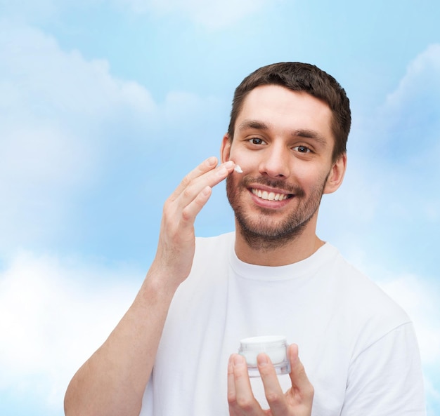 health and beauty concept - beautiful smiling man with jar of cream applying cream